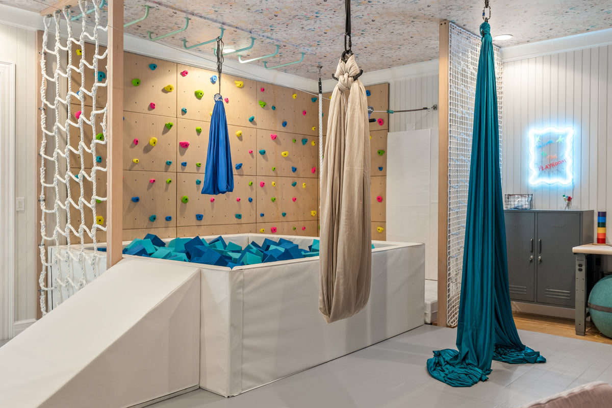 An indoor playroom featuring a large white climbing wall with a foam pit, colorful climbing holds, hanging blue aerial hammocks, and a teal fabric sling suspended in the room.